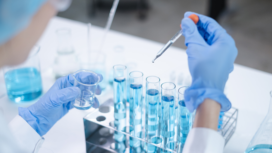 Osoba u laboratorijskoj opremi i plavim rukavicama drži epruvetu i pipetu, dok dodaje tečnost u red epruveta sa plavom tečnošću. U pozadini se vide različite laboratorijske posude.