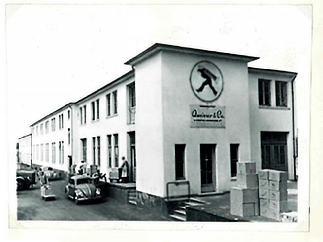 Na slici je zgrada sa natpisom "Quinever & Co.", sa stilizovanom slikom muškarca koji nosi teret. Ispred zgrade su parkirani automobili iz prošlih decenija, a pored njih nekoliko ljudi i kutije. Zgrada ima prozore sa roletnama.