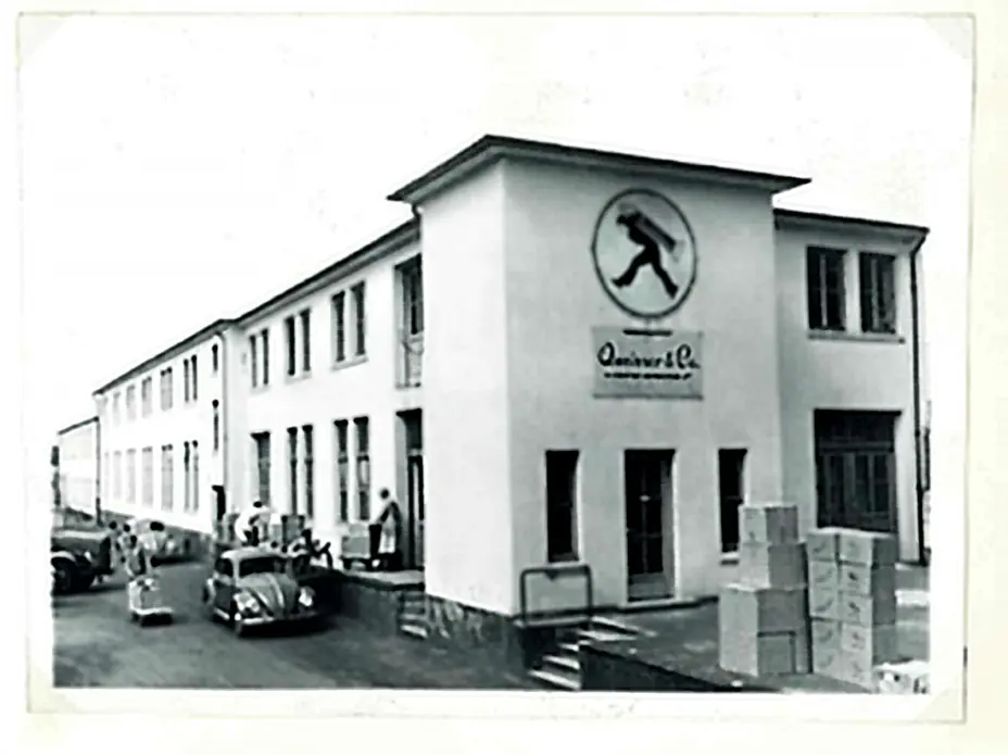 Na slici je zgrada sa natpisom "Quinever & Co.", sa stilizovanom slikom muškarca koji nosi teret. Ispred zgrade su parkirani automobili iz prošlih decenija, a pored njih nekoliko ljudi i kutije. Zgrada ima prozore sa roletnama.