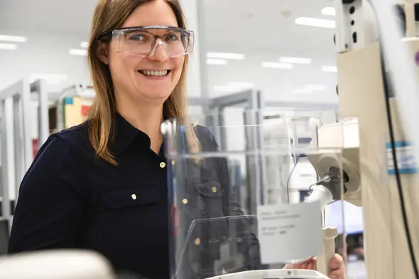 Žena sa smeškom nosi zaštitne naočare i stoji pored laboratorijske opreme. U pozadini se vidi radni prostor sa više laboratorijskih mesta.