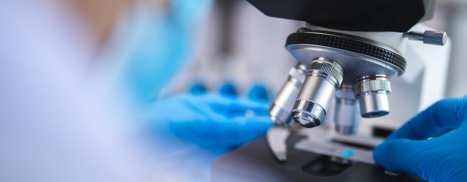 Laboratorijski mikroskop sa plavim rukavicama koje ga koriste. Fokus je na mikroskopu i na pripremi uzorka ispod objektiva. U pozadini se može videti zamućen lik osobe koja radi u laboratoriji.