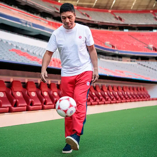 Na fotografiji je muškarac u sportskom izgledu kako igra fudbal na travnatoj površini unutar fudbalskog stadiona. Obučen je u belu majicu i crvene trenerke. U pozadini se vide tribine stadiona sa praznim sedištima.