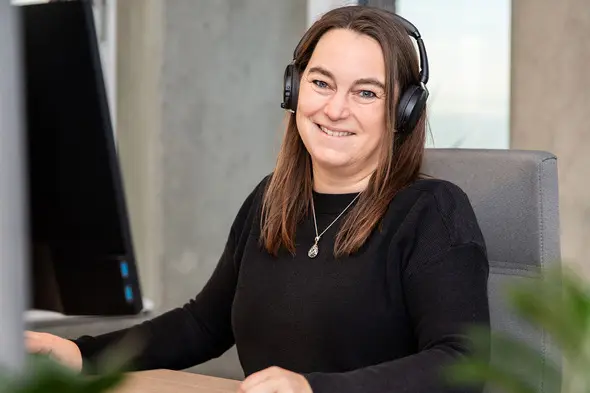 Žena sa slušalicama sedi za radnim stolom i osmehuje se. U pozadini se vidi računar. Ambijent deluje moderan i prijatan.
