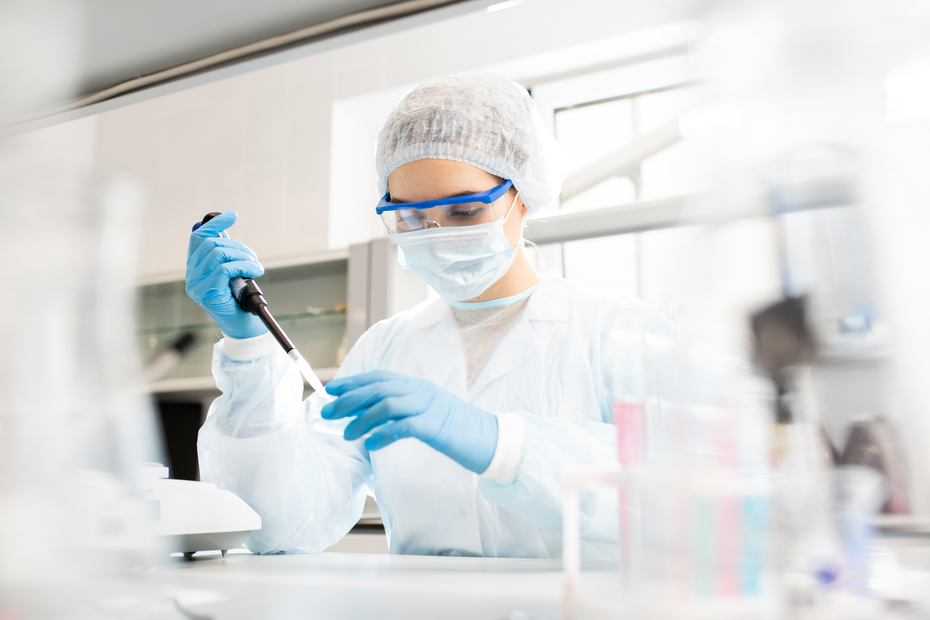 Laborant u zaštitnoj opremi koristi pipetu u laboratoriji. Nosite masku, rukavice i zaštitne naočare dok radi sa hemijskim supstancama na stolu u pozadini.