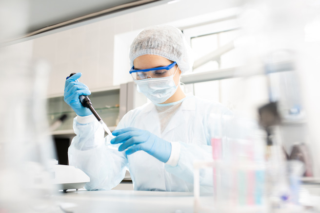 Laborant u zaštitnoj opremi koristi pipetu u laboratoriji. Nosite masku, rukavice i zaštitne naočare dok radi sa hemijskim supstancama na stolu u pozadini.
