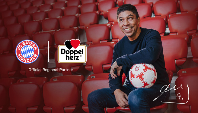 Na slici se nalazi nasmejani muškarac koji sedi na stadionu sa praznim redovima. Drži fudbalsku loptu u rukama, a iza njega su logotipi fudbalskog kluba Bayern Minhen i kompanije Doppelherz.