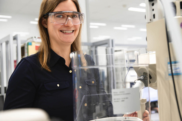 Žena sa smeškom nosi zaštitne naočare i stoji pored laboratorijske opreme. U pozadini se vidi radni prostor sa više laboratorijskih mesta.